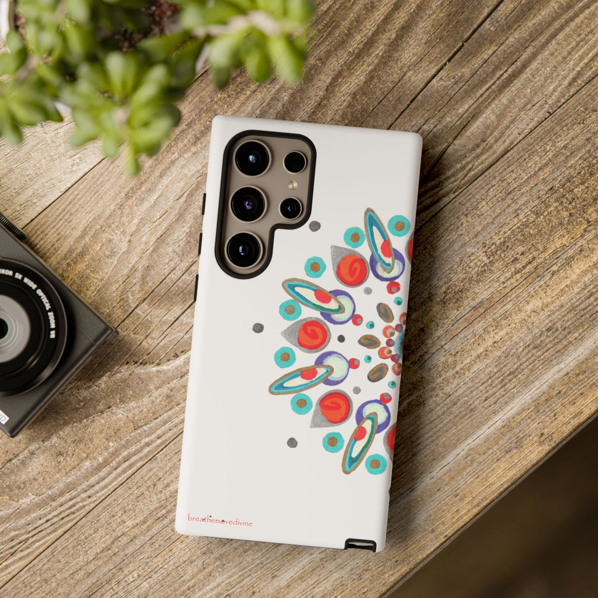 Phone case with colorful design on a wooden surface next to a camera and plant