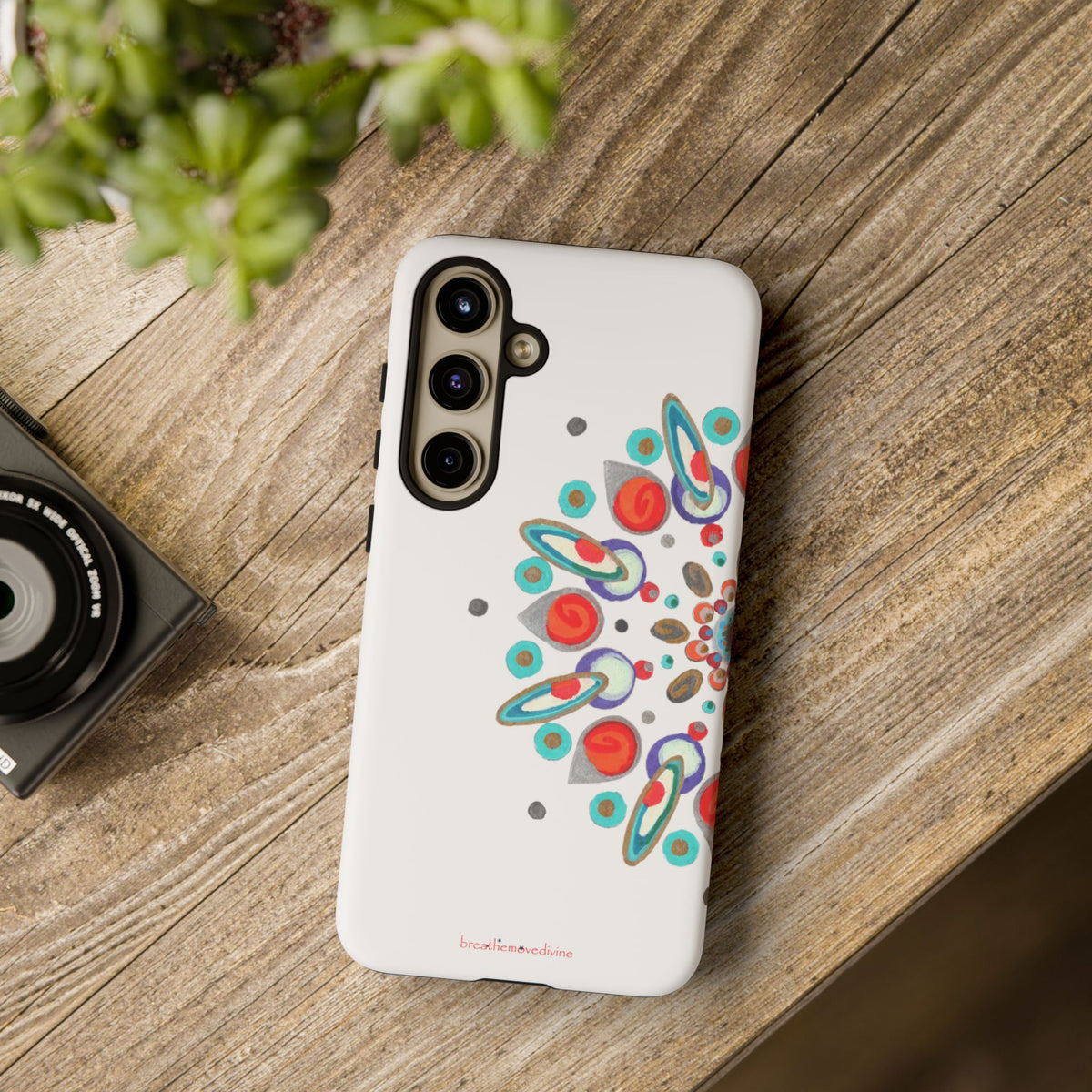 Phone case with colorful design on a wooden surface next to a camera and plant