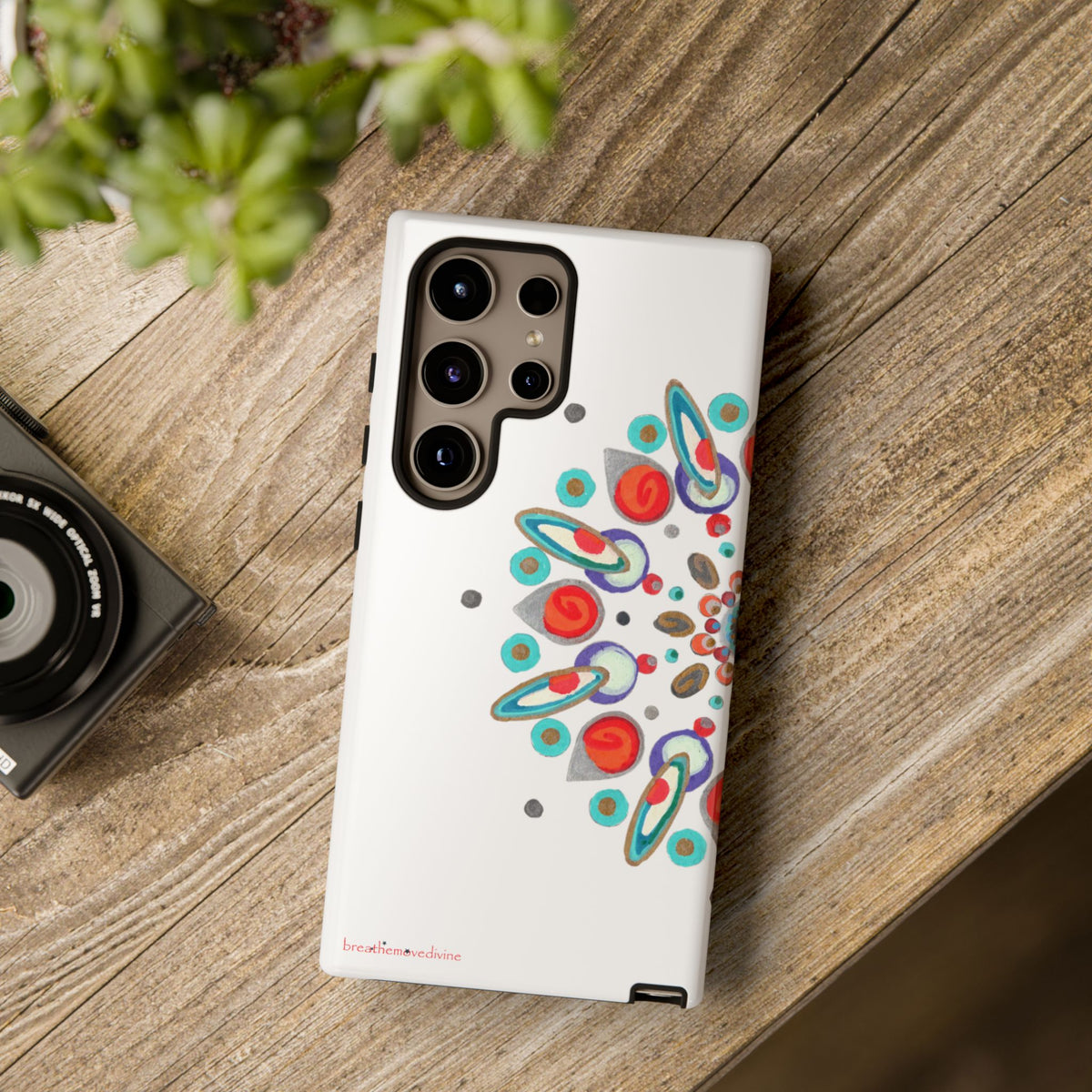Phone case with colorful design on a wooden surface next to a camera and plant
