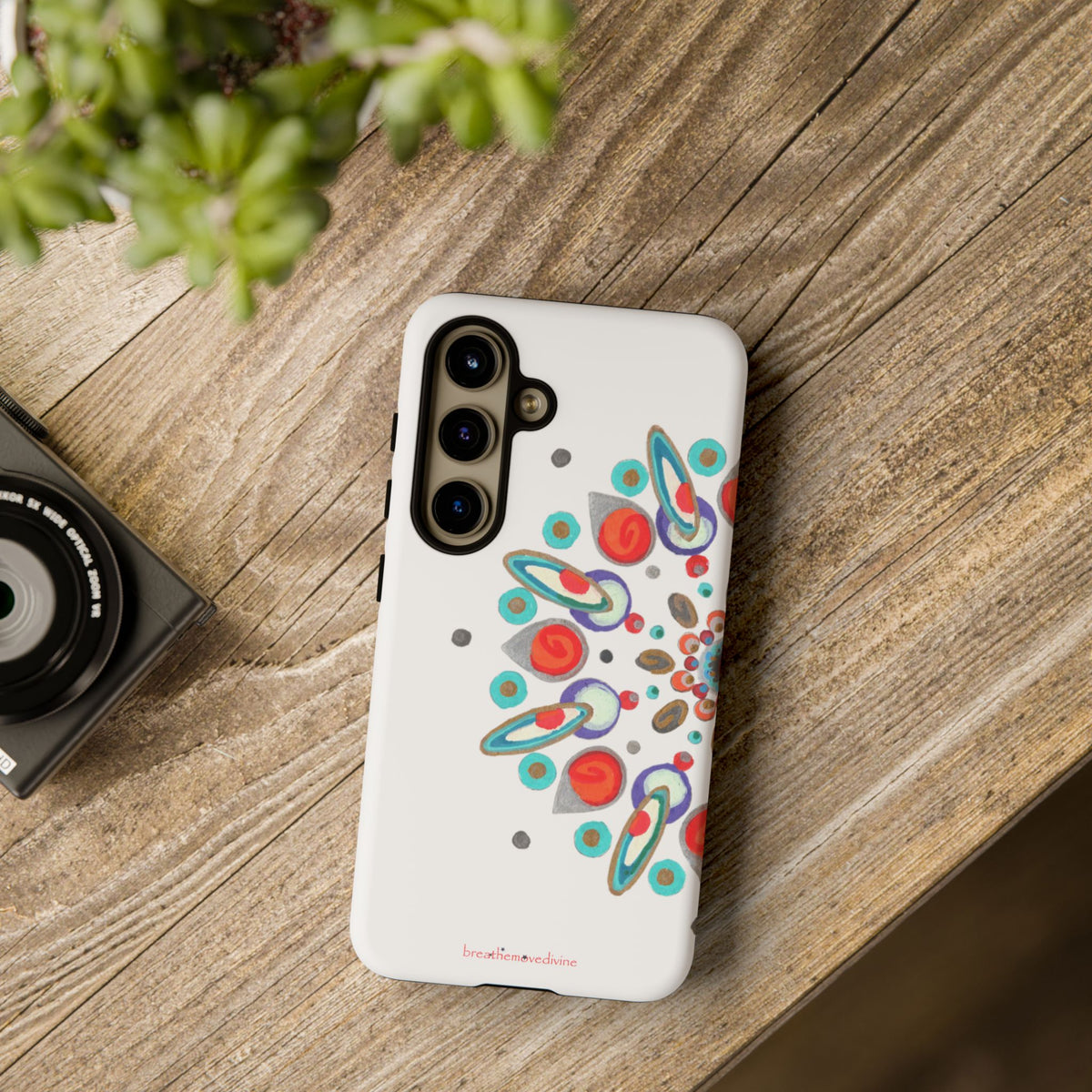 Phone case with colorful abstract design on a wooden surface next to a camera and plant.